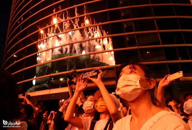 در حالی که مردم مراسم افتتاحیه را از بیرون ورزشگاه المپیک در توکیو ، ژاپن ، 23 جولای 2021 مشاهده می کنند ، آتش بازی در یک ساختمان منعکس می شود.